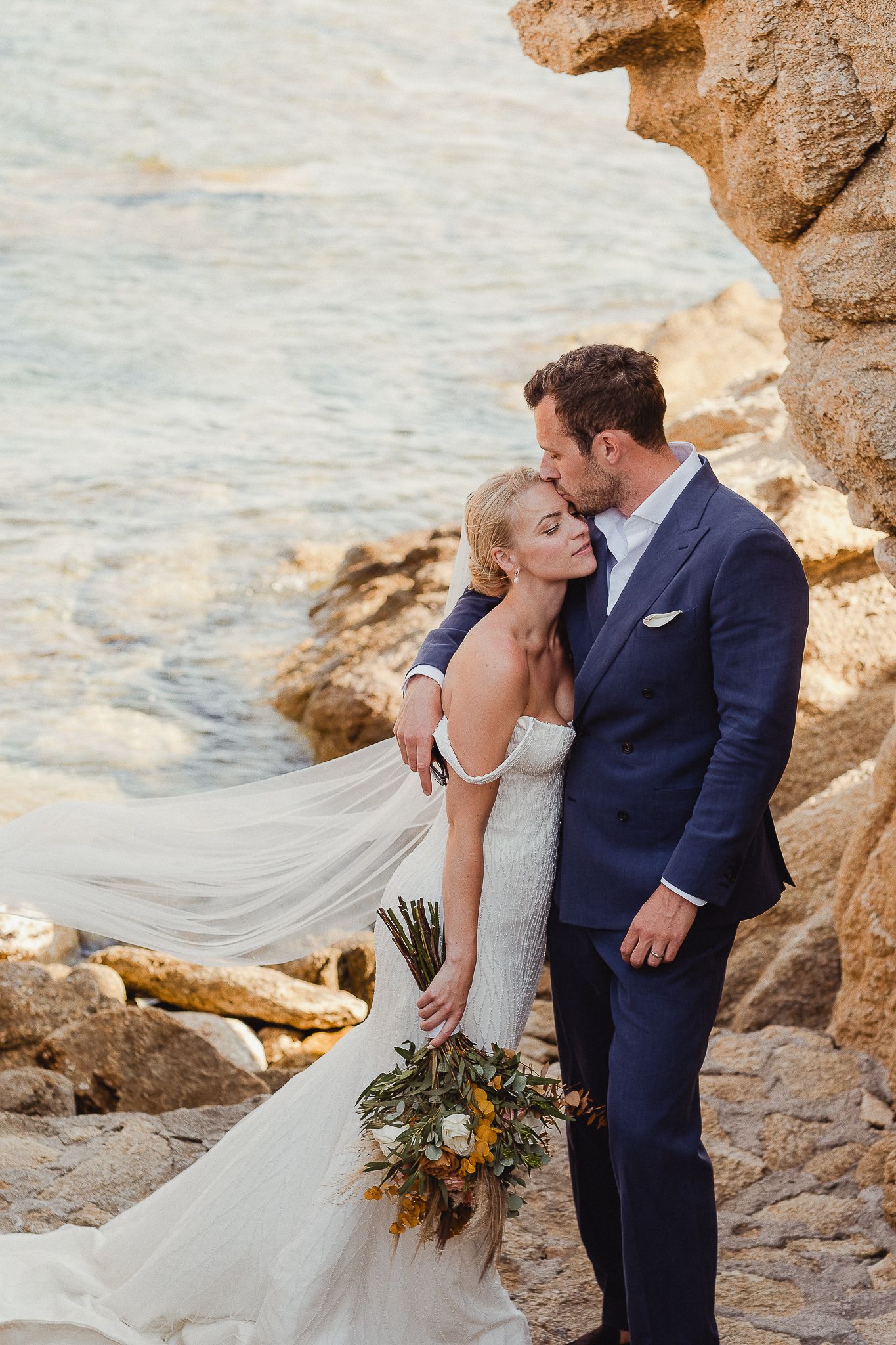 A romantic coastal wedding photoshoot shows sweet moments between a couple by rocky cliffs at sunset.