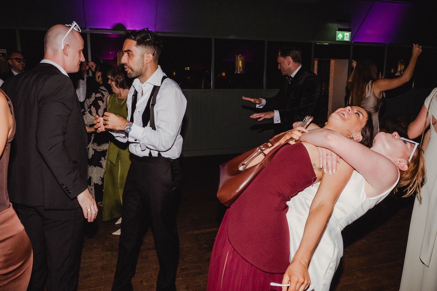 People dancing and socializing at a nighttime wedding reception with purple lighting in the background.