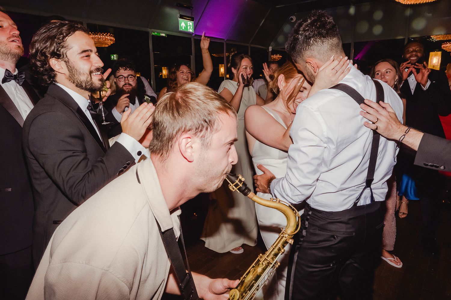 Wedding guests dancing and celebrating on a crowded dance floor with colorful purple lighting in the background.