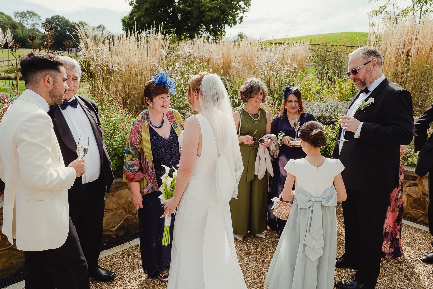 A wedding party gathers in a garden setting with tall grasses and natural landscaping in the background.
