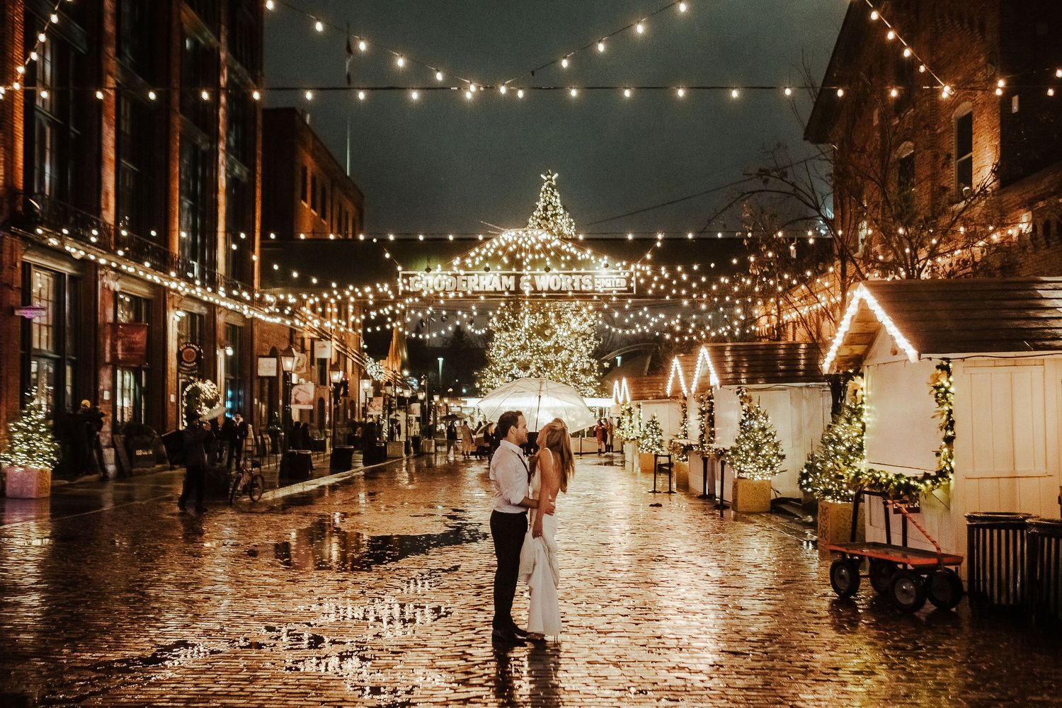A couple stands in a romantic rainy evening under string lights at a festive outdoor market decorated for the holidays.