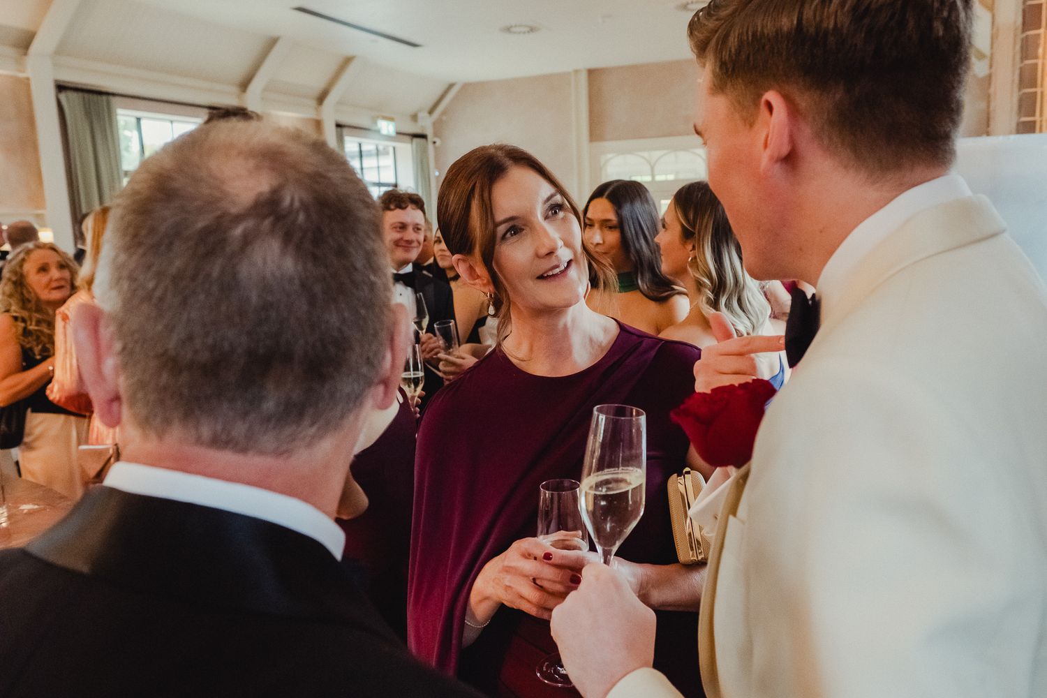 Wedding guests mingle and chat while holding champagne glasses at an elegant indoor reception.