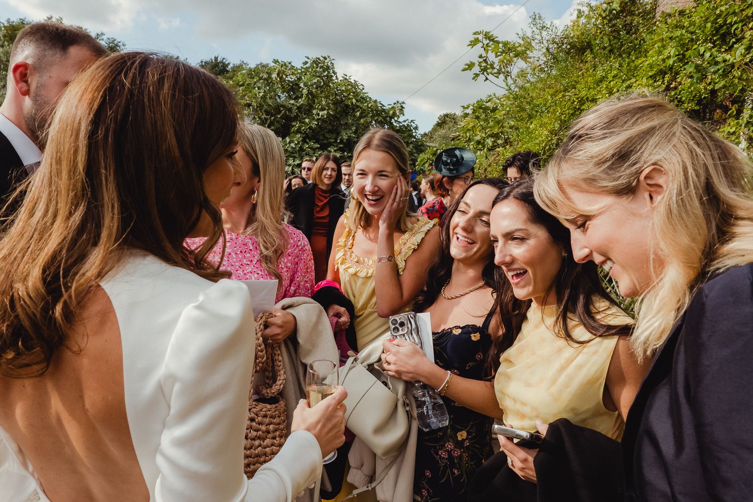 A group of wedding guests laugh together while gathered outdoors during a celebration.