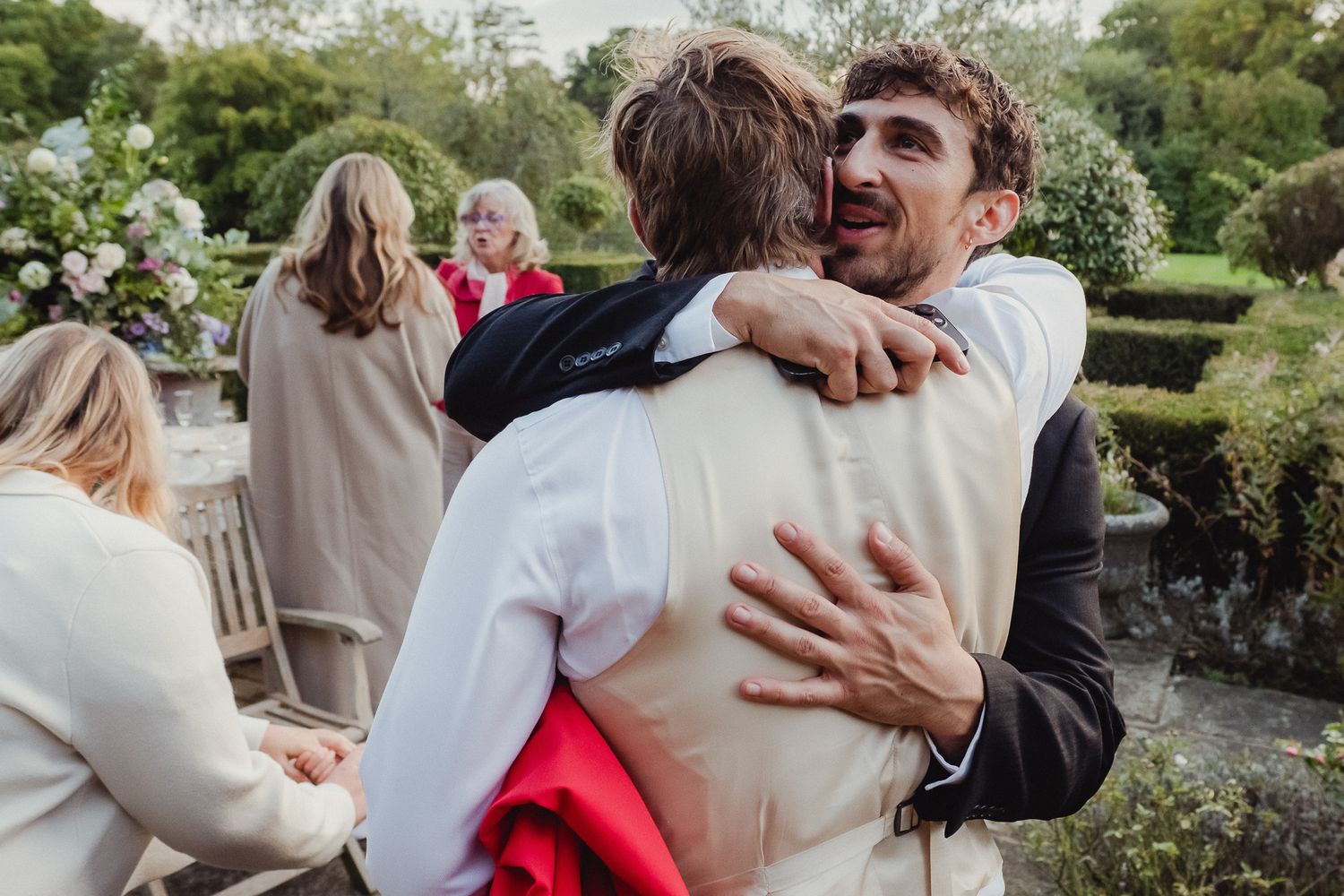 A couple shares an intimate embrace in a garden setting while wearing light colored clothing.