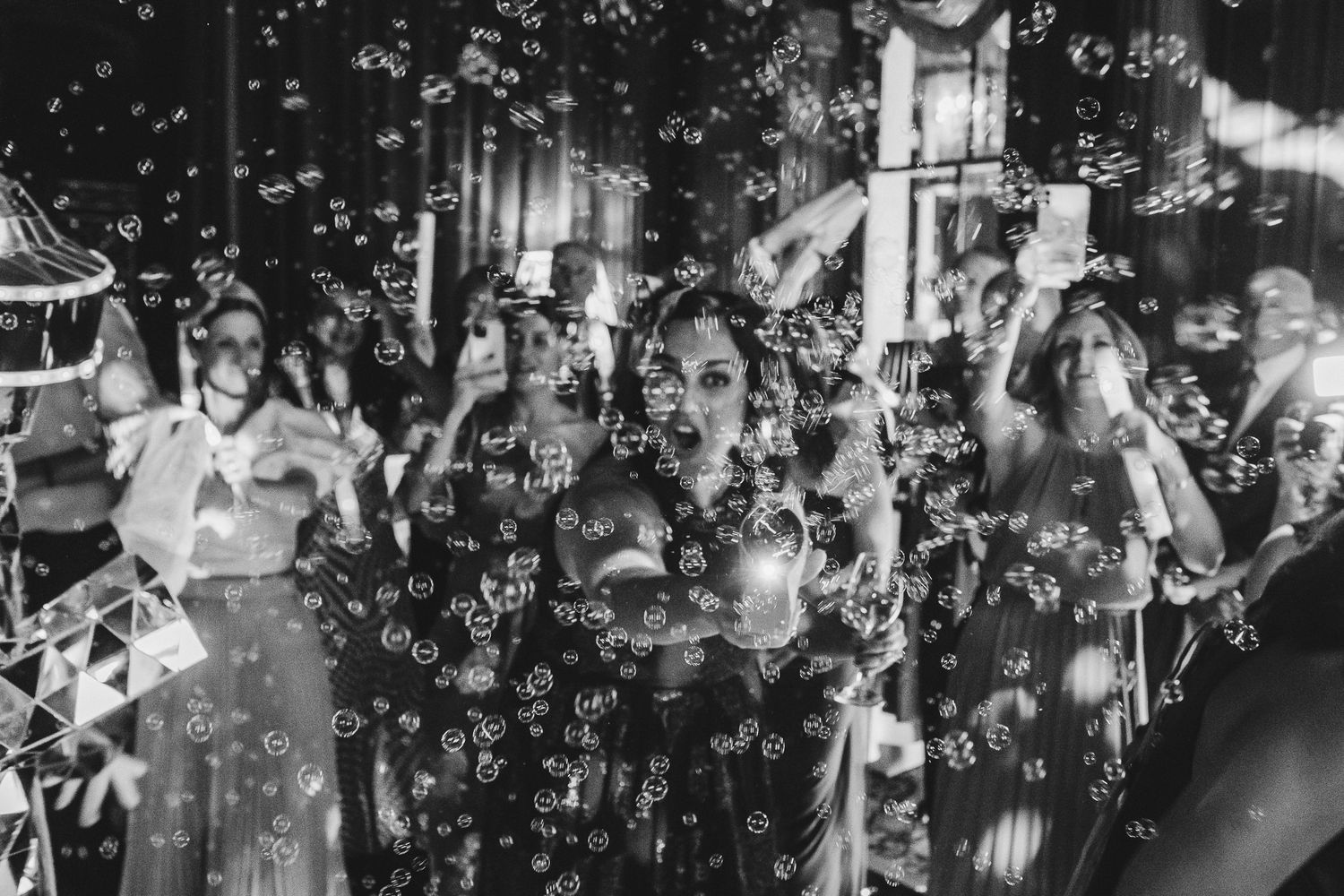 Wedding guests blow bubbles and celebrate as newlyweds walk through a joyful crowd at an outdoor reception.