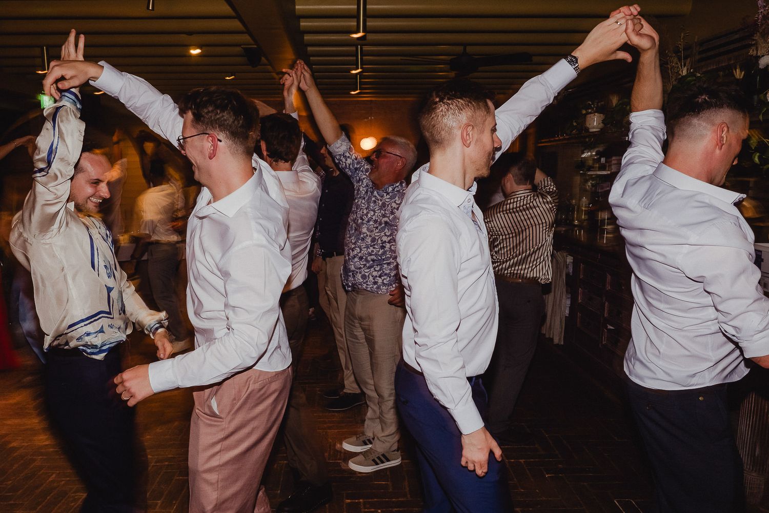 Wedding guests dance together with raised arms during a lively reception celebration on the dance floor.