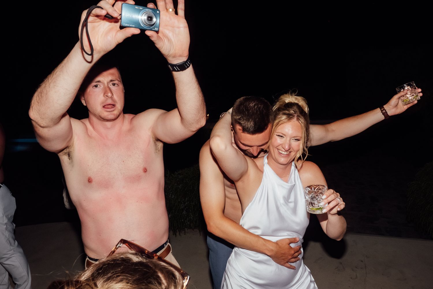 Shirtless man photographs couple dancing at lively outdoor night party celebration.