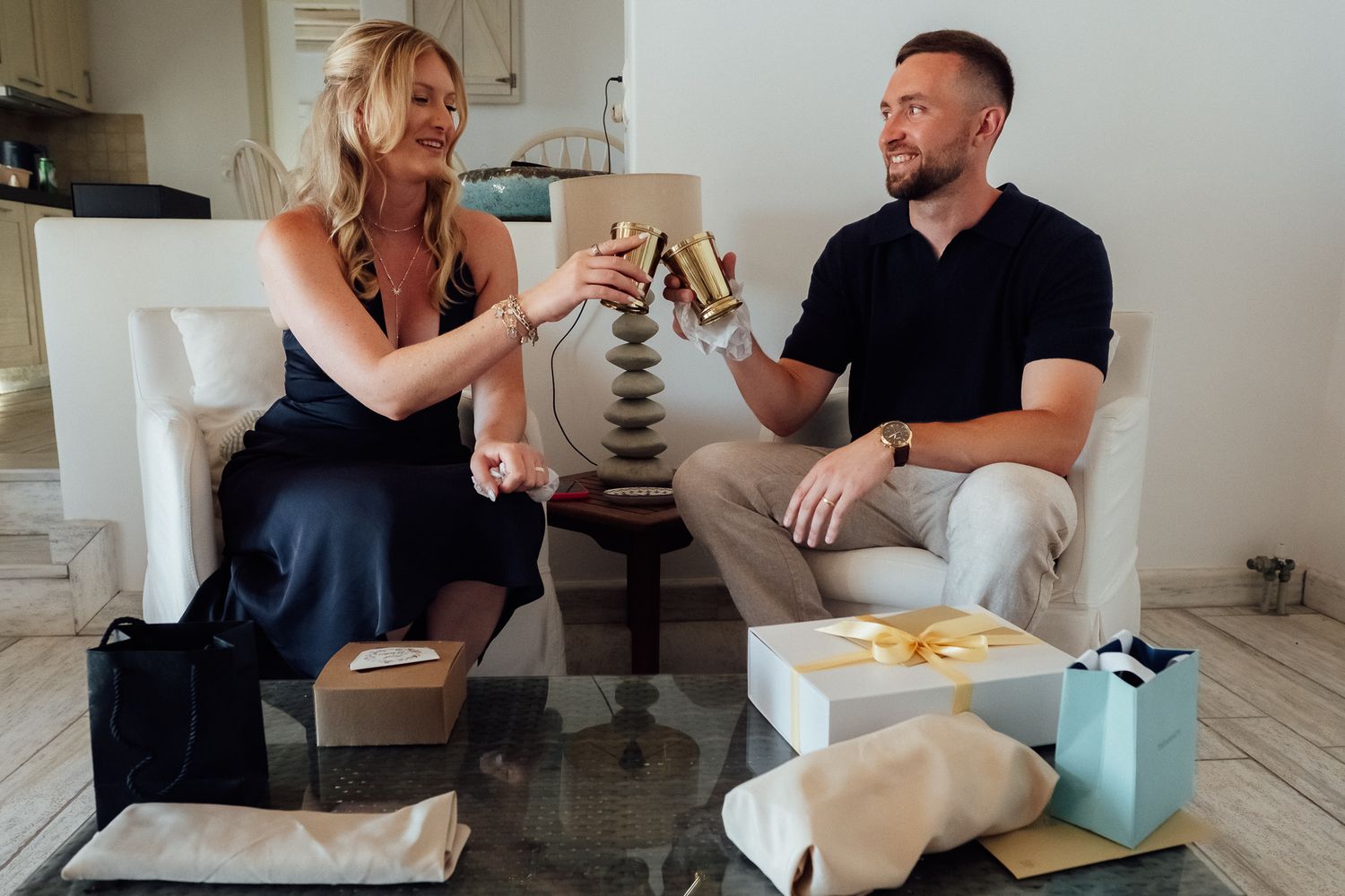 A couple toasting with gold champagne flutes surrounded by gift boxes in a bright living room setting.