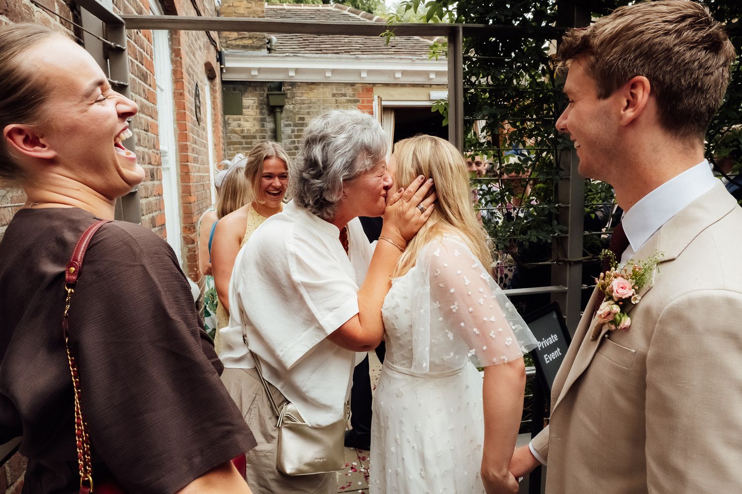 Elderly woman kissing bride in white dress at outdoor wedding celebration while groom and guests smile nearby.