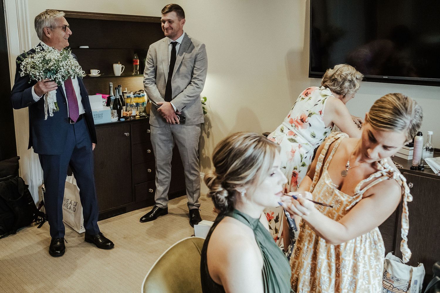 People gather in a room during wedding preparations as someone helps with a floral dress.