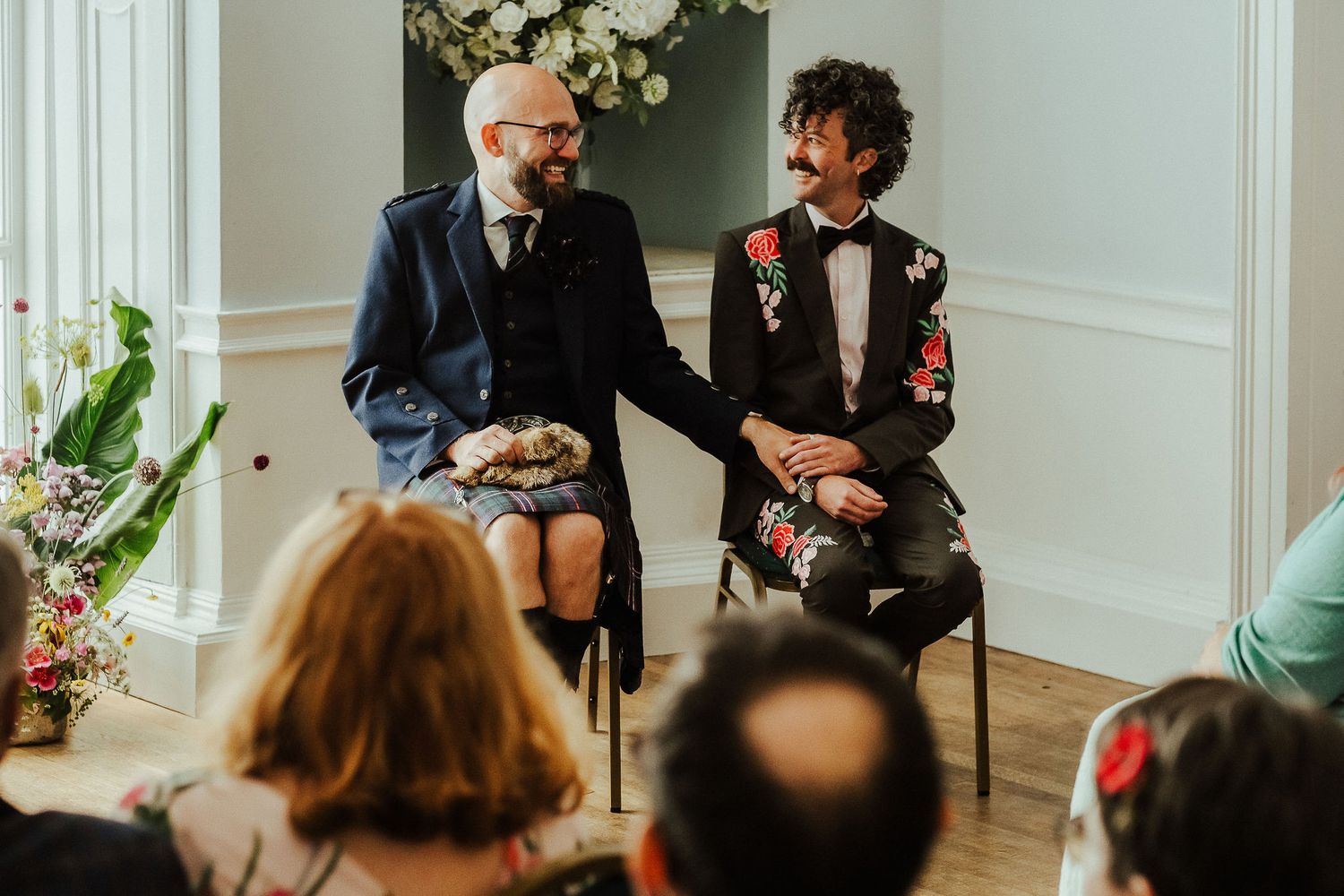 Two grooms in a navy suit and a floral embroidered jacket hold hands and smile during an intimate wedding ceremony as guests watch.