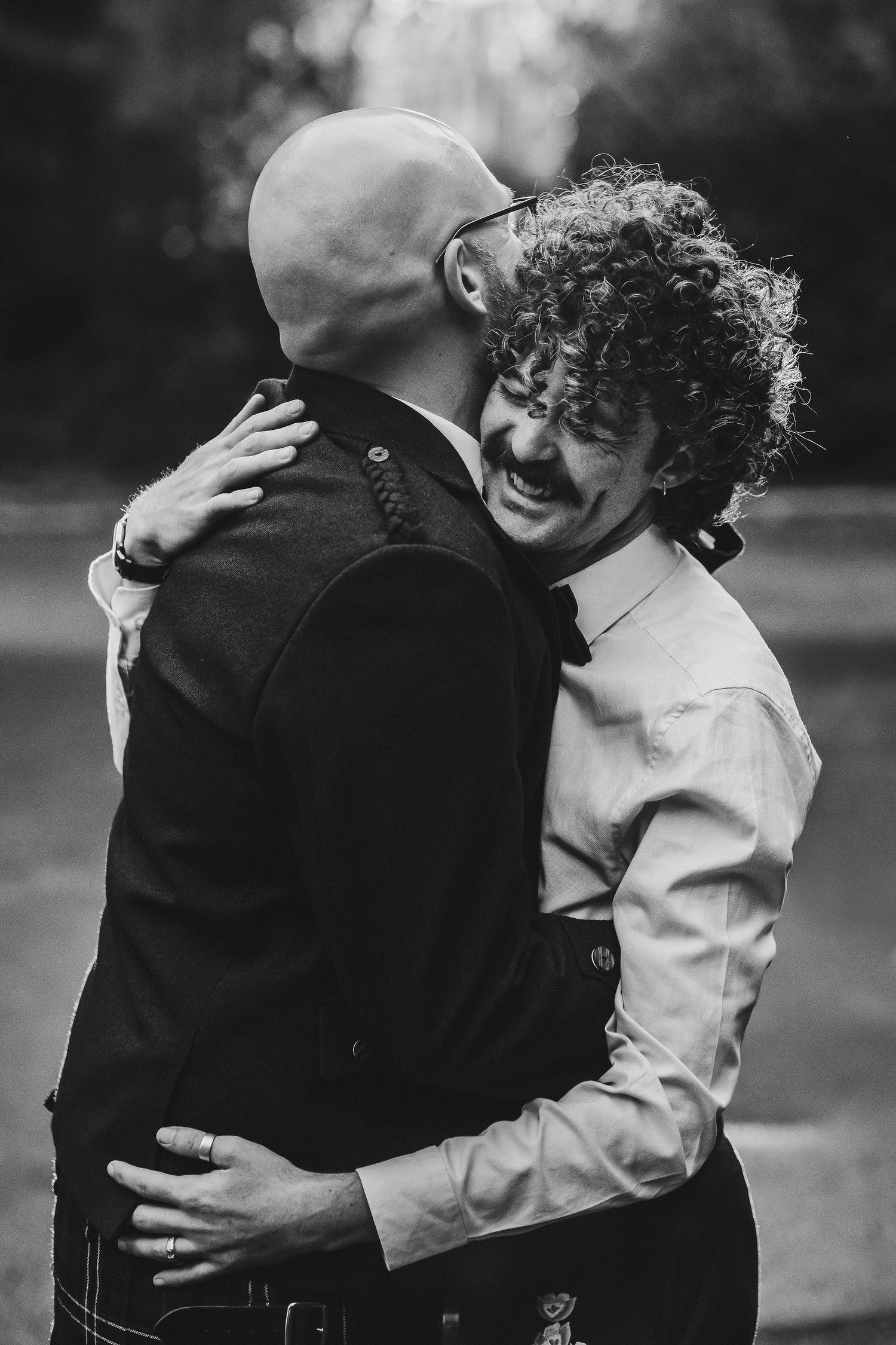 Black and white photo of two men sharing a joyful embrace outdoors, one with curly hair and a mustache smiling in a bow tie.