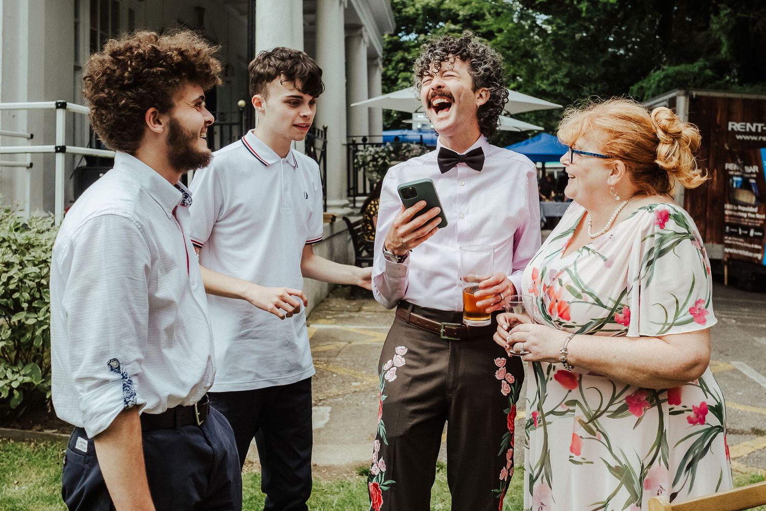 Four people laugh and chat outdoors at a casual gathering, including a man in a bow tie holding a phone and a woman in a floral dress.