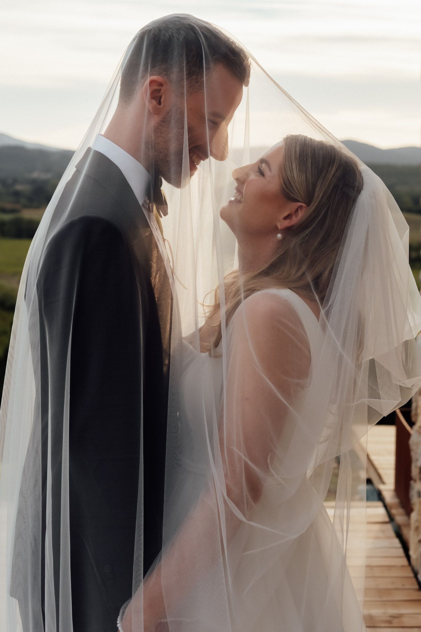 Bride and groom smile at each other beneath a sheer wedding veil with rolling hills in the background.