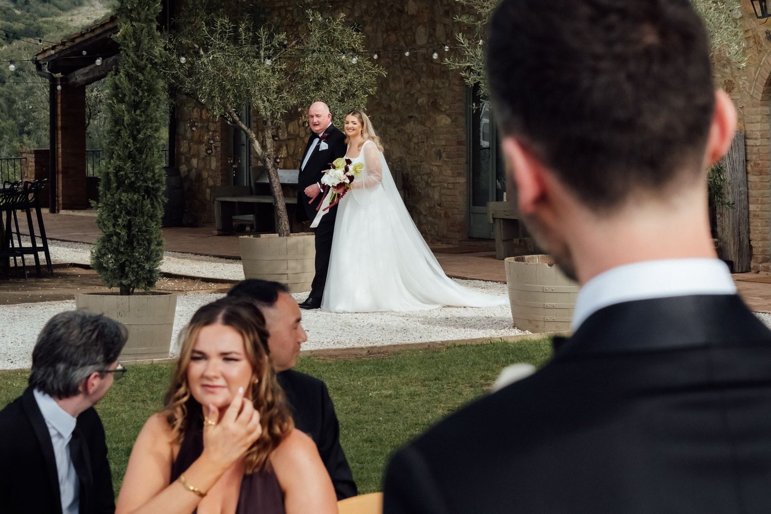 Bride in a white gown and groom in a dark suit stand outdoors at a rustic wedding ceremony as emotional guests watch from the foreground.
