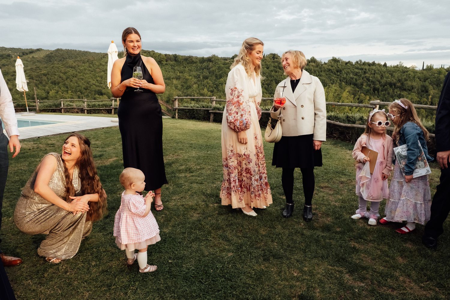 Women and young children in dresses gather and laugh on a green lawn at an outdoor wedding celebration with hills in the background.