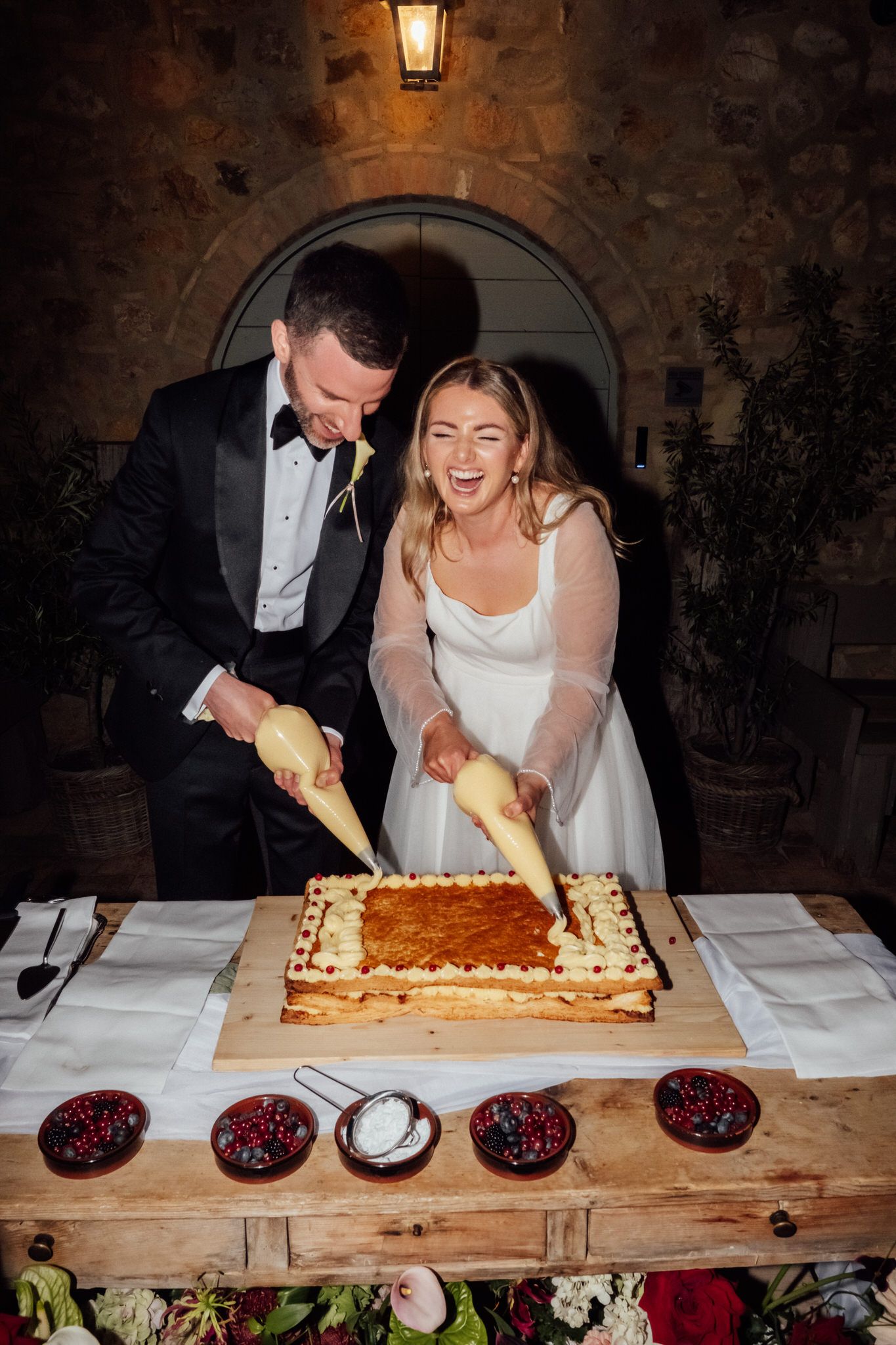 Smiling bride and groom in formal wedding attire pipe cream onto a large rustic cake on a wooden table in a candlelit stone venue.