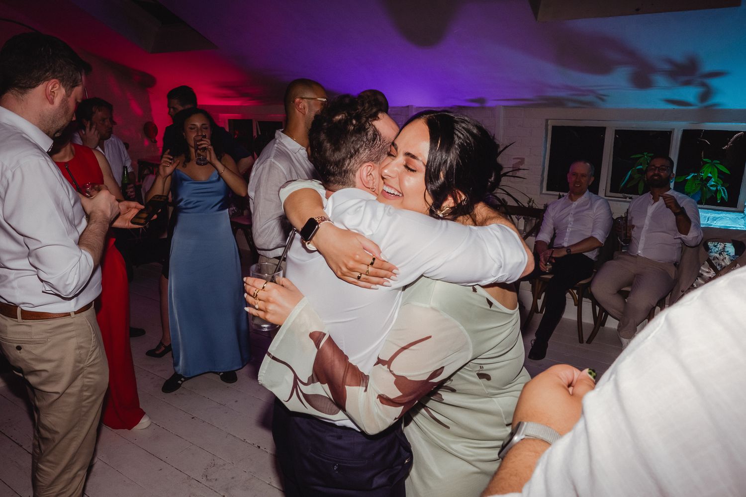 Wedding reception guests embrace on the dance floor under colorful purple and blue lighting.