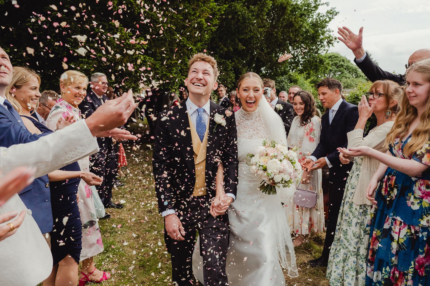 Wedding guests celebrate by throwing confetti over the happy couple during an outdoor countryside celebration.