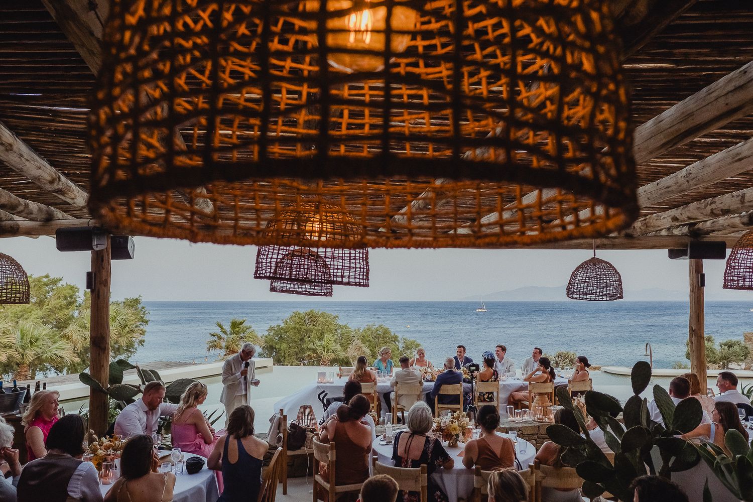 A scenic thatched-roof outdoor beach restaurant overlooking the ocean with guests dining under natural shade.