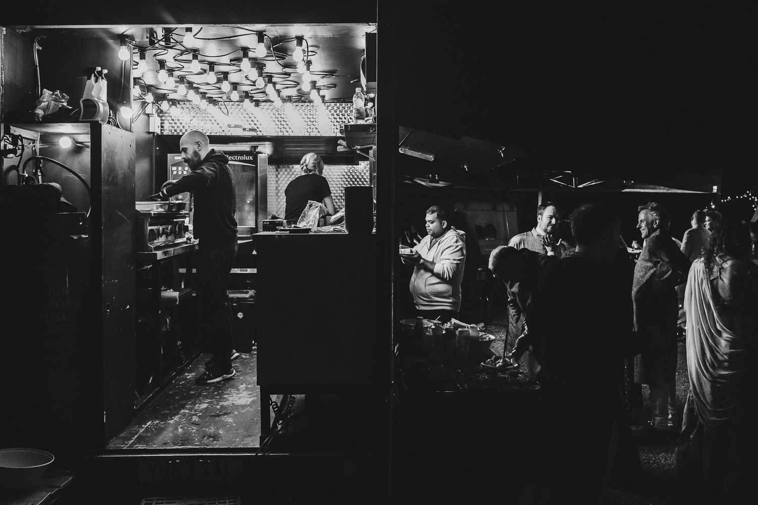 Atmospheric black and white nighttime scene of people gathered outside a lit venue.