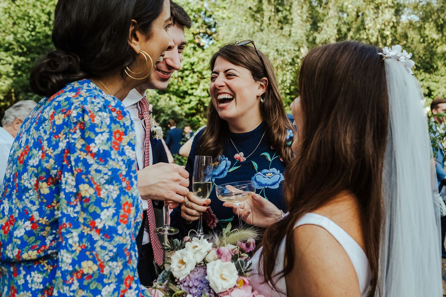 Wedding guests share joyful laughter while holding drinks at an outdoor celebration.