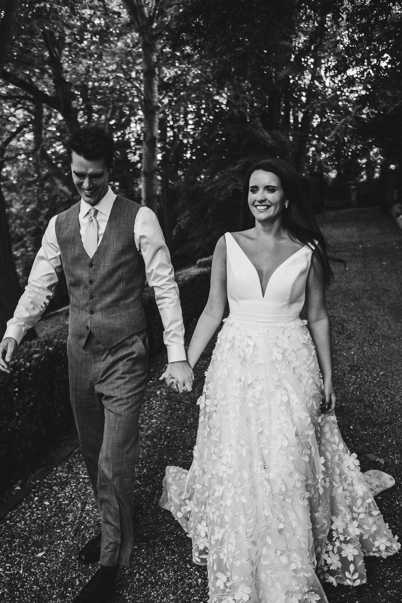 Bride and groom walk together on gravel path through wooded area.