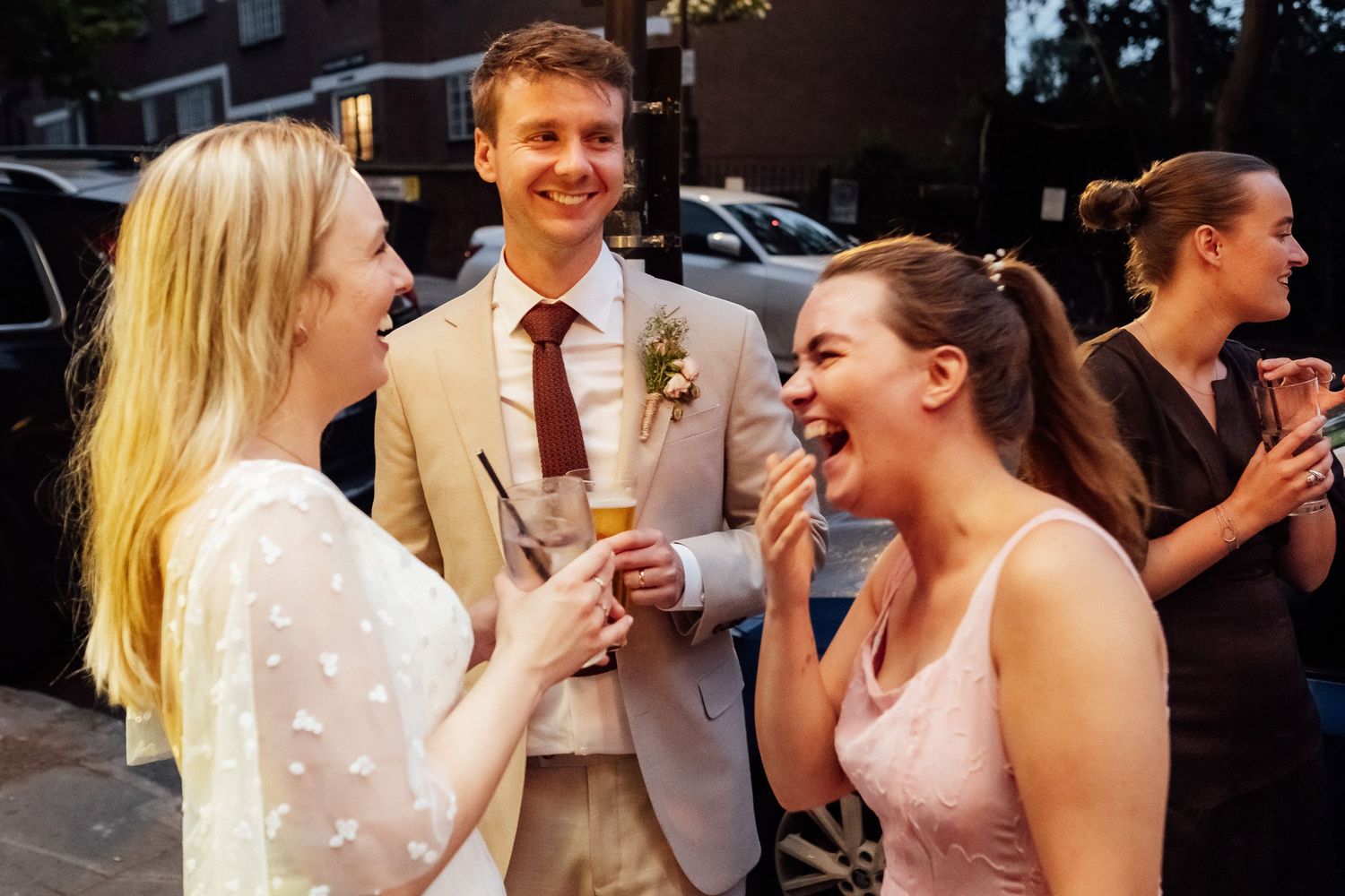 Group of friends laughing and chatting outdoors at an evening event, with a man in a tan blazer and boutonniere holding a drink.