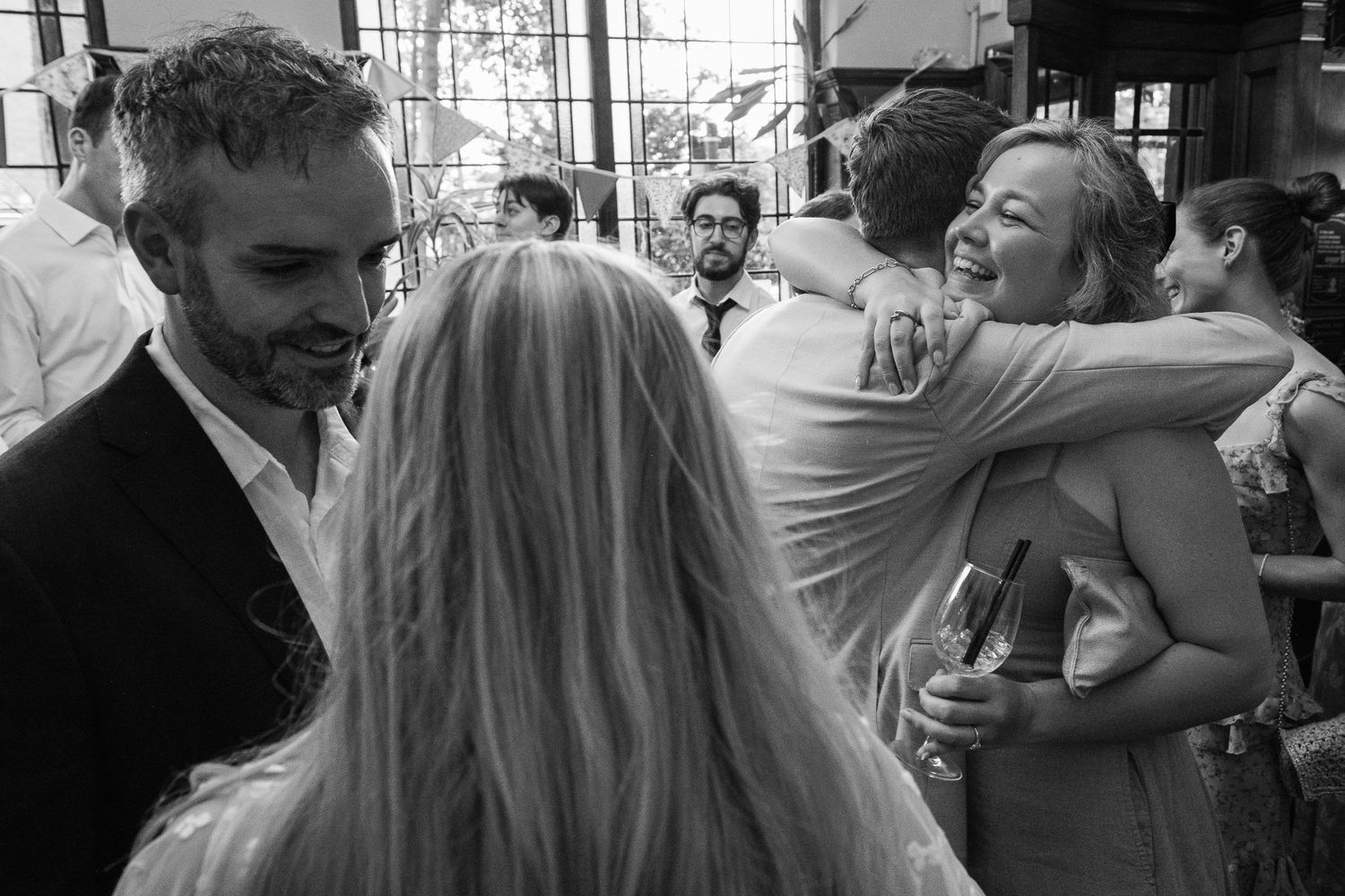 Black and white photo of guests embracing and smiling at a lively indoor celebration, with a man in a blazer looking on.
