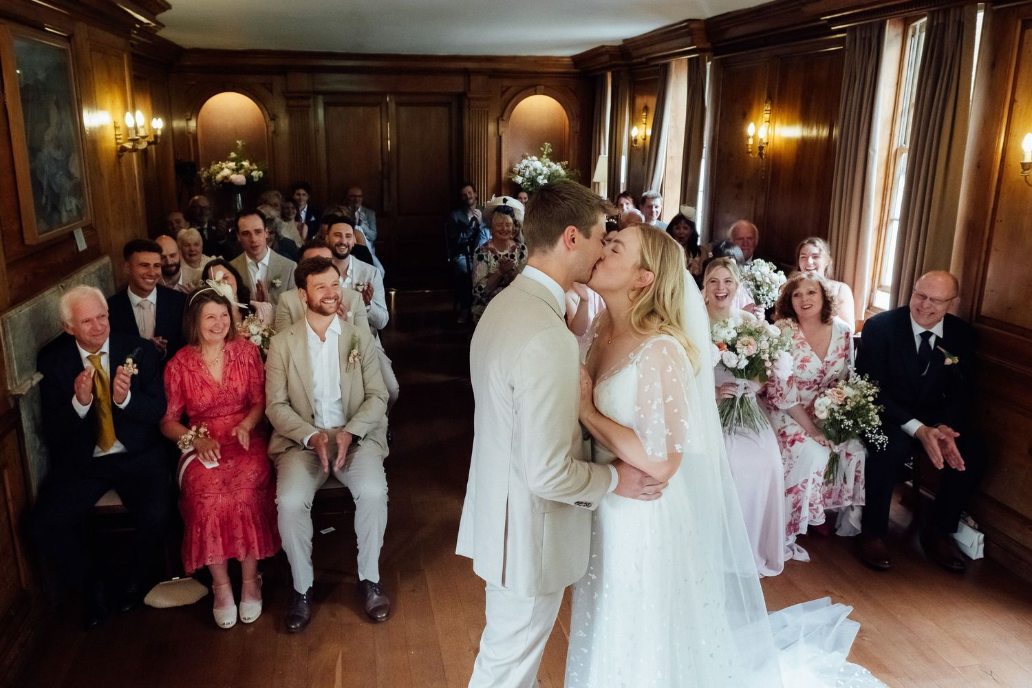 Bride and groom share a kiss during their wedding ceremony in a wood-paneled room as seated guests watch and smile.