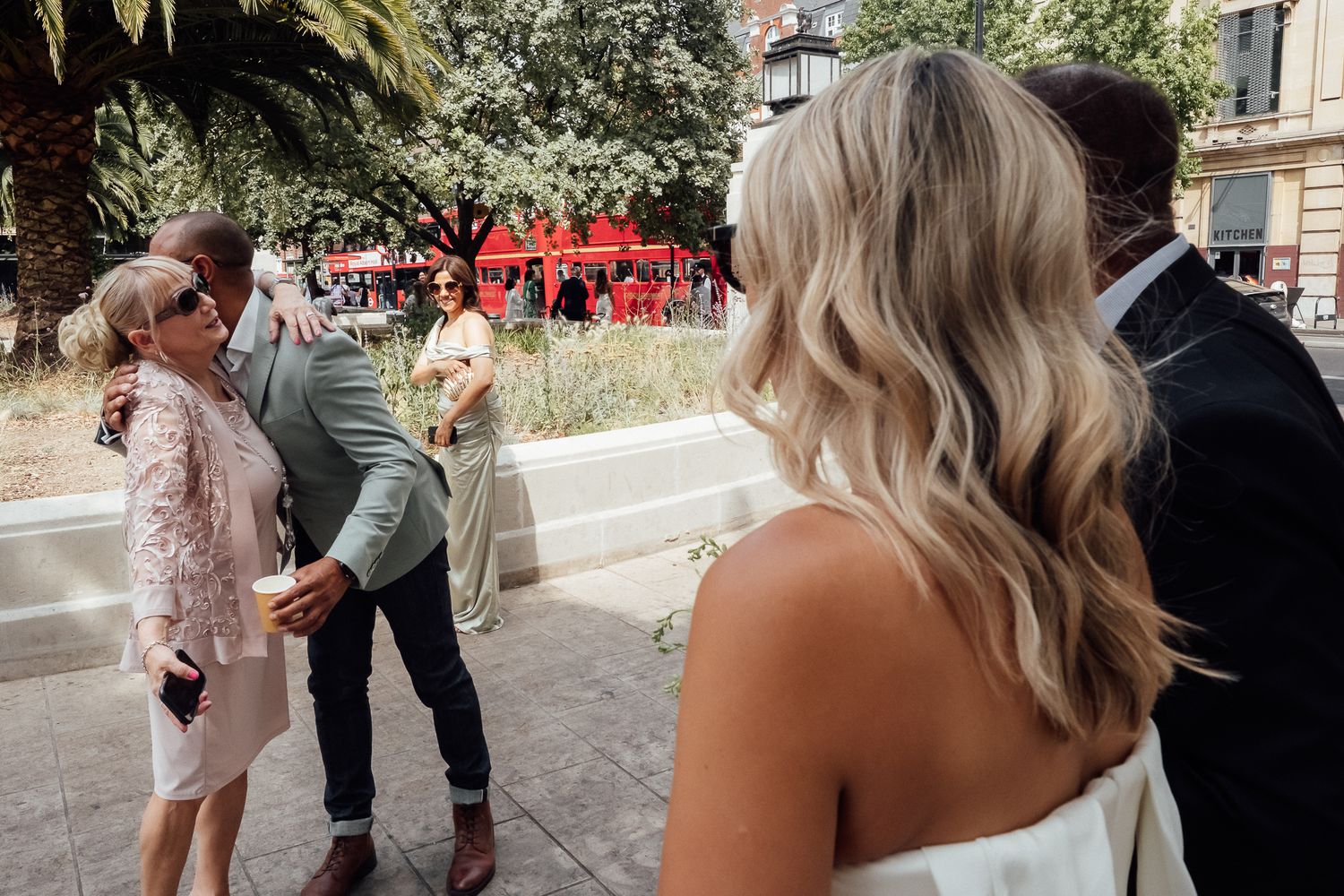Blonde woman in a white dress watches guests greet each other with hugs at an outdoor wedding gathering near palm trees and a plaza.