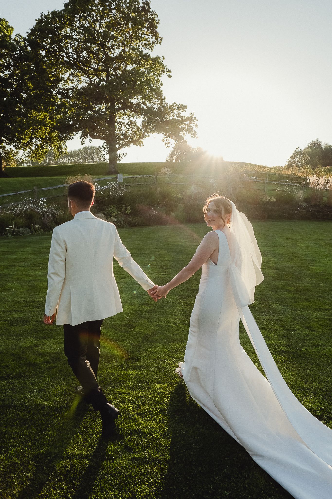 A couple in wedding attire runs hand in hand through a sunlit meadow at golden hour.