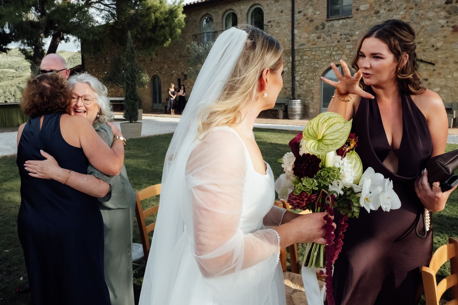 Bride in a long veil holds a lush bouquet at an outdoor Italian wedding while guests hug and a woman in a burgundy dress looks on.