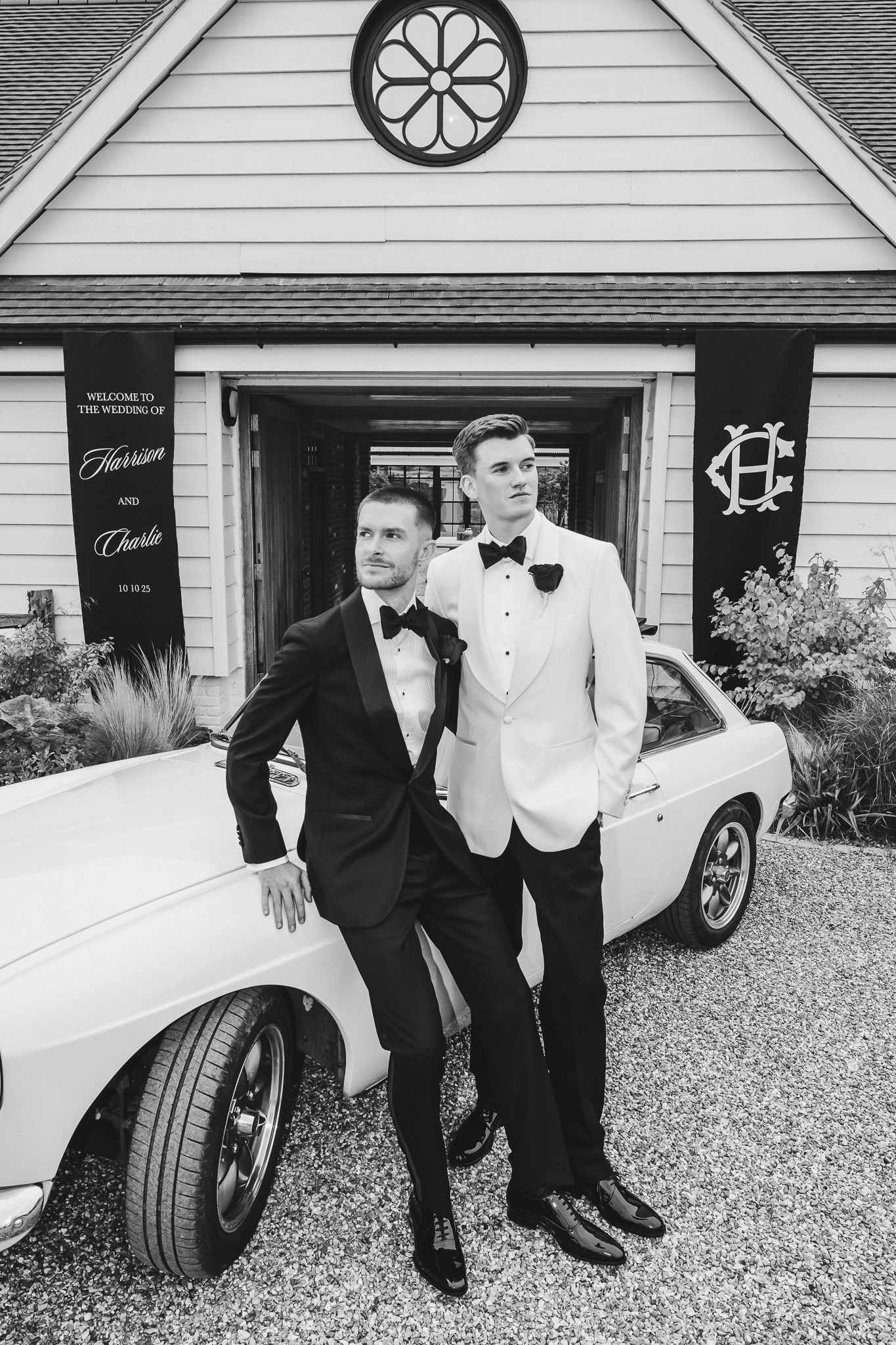 Black and white photo of groomsmen in tuxedos posing casually beside a classic white car.