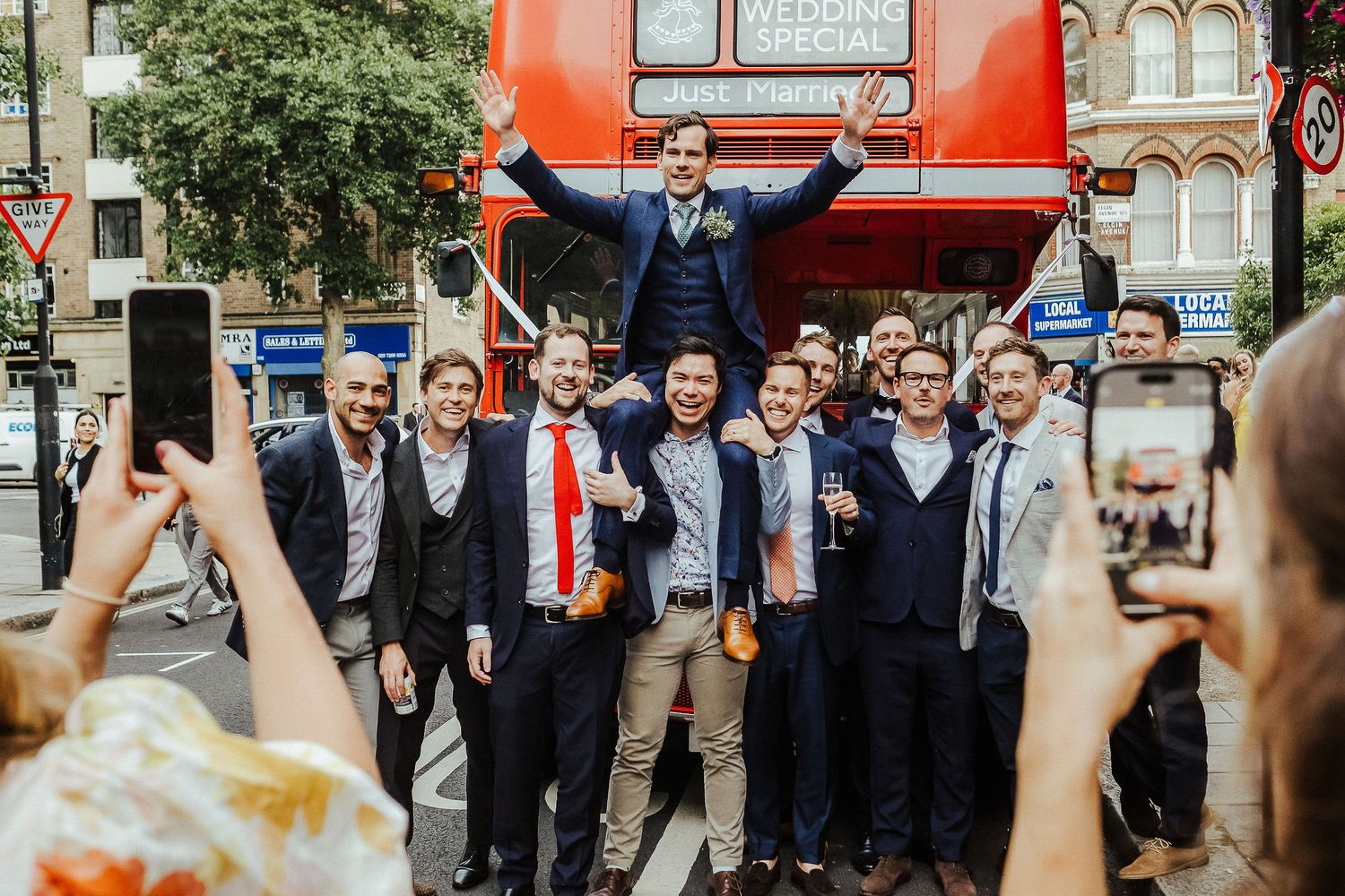 Wedding party poses in front of a red double-decker bus on a London street.