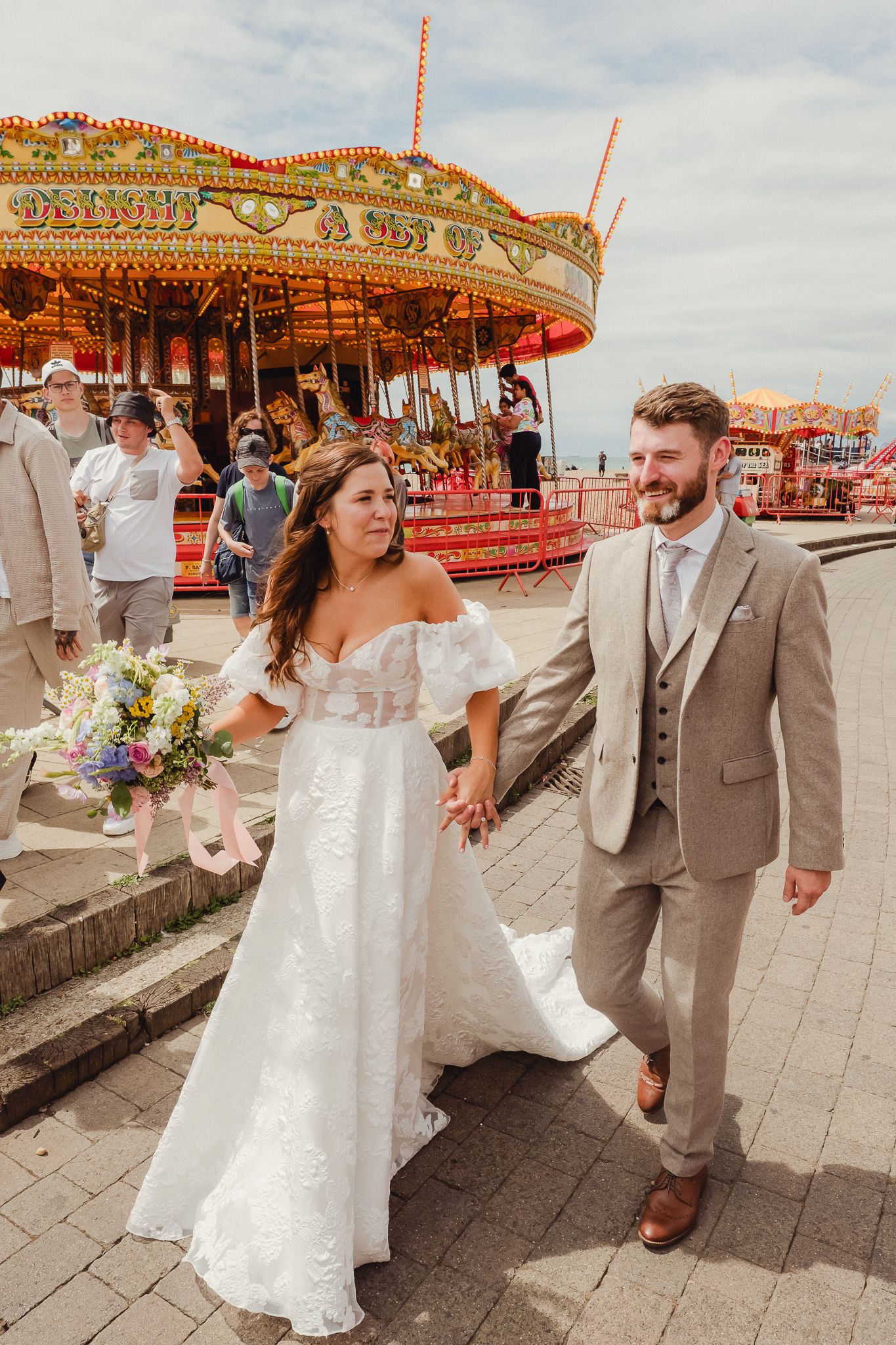 A wedding couple poses playfully in front of a vintage carousel at an amusement park setting.