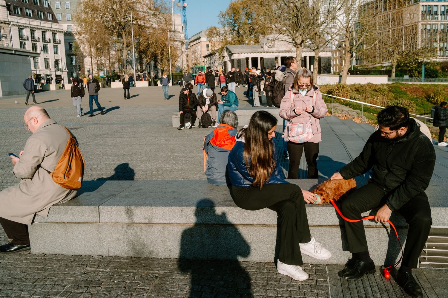 People relaxing and socializing in an urban plaza on a sunny autumn day with fallen leaves and city buildings.