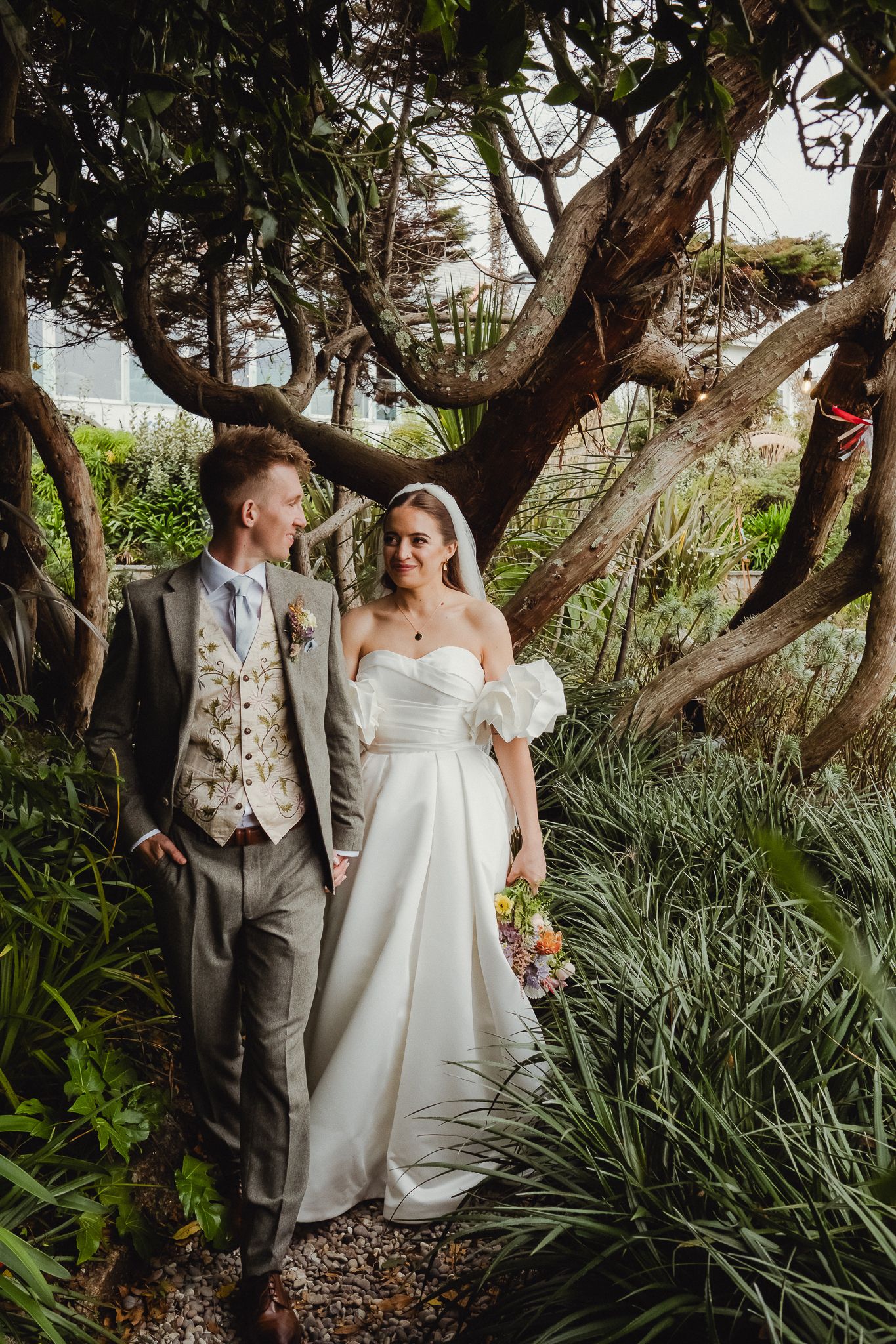 A couple in wedding attire share intimate moments among lush tropical foliage in a garden setting.