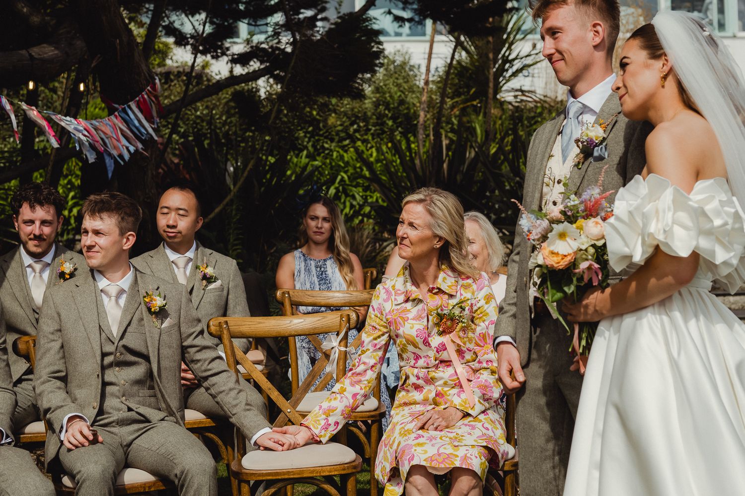 Wedding party laughs together during outdoor celebration with bridesmaids in floral dresses and groomsmen in gray suits.