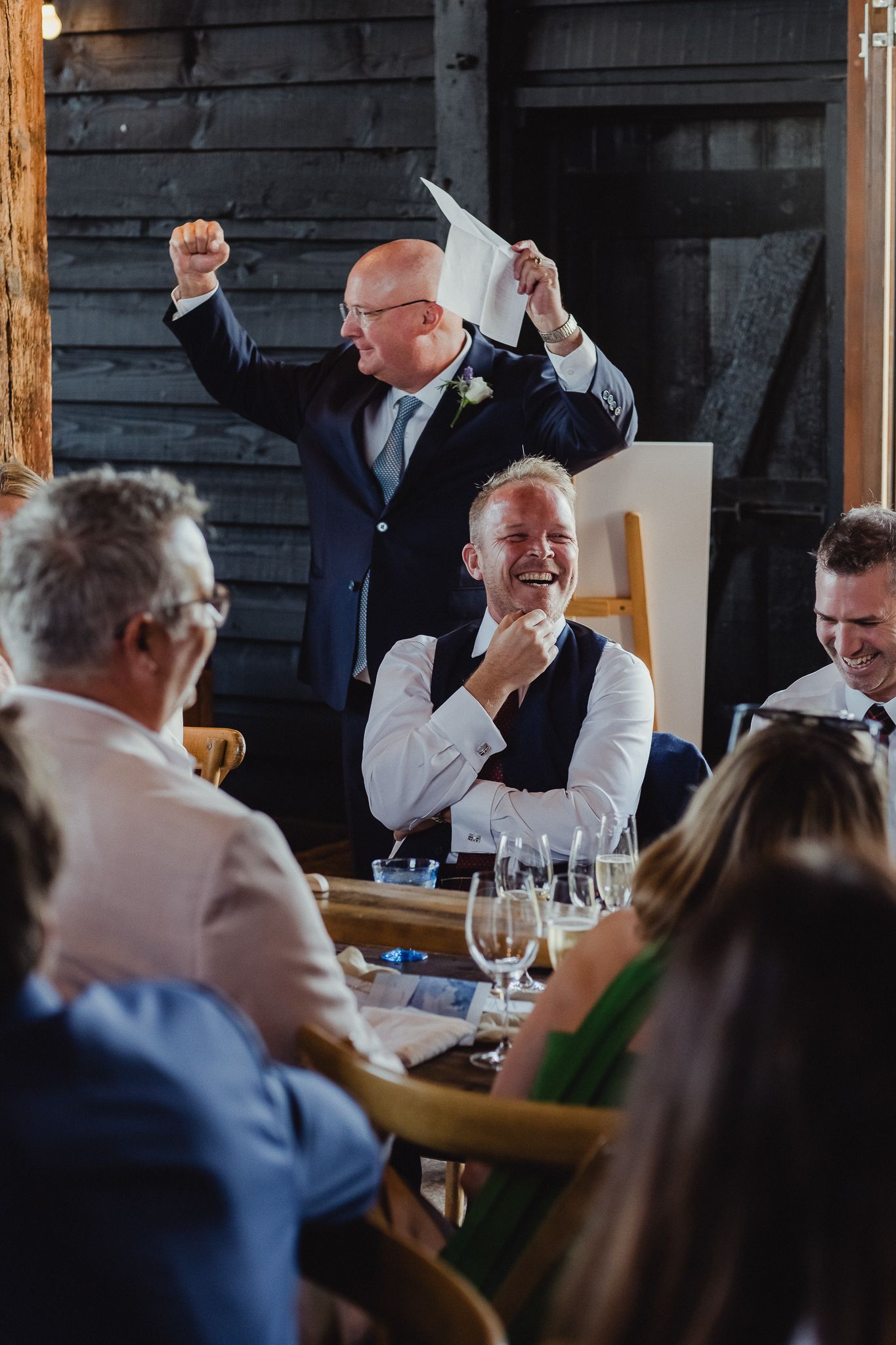 Wedding guests celebrate and raise their hands in joy during a reception dinner at a rustic venue.