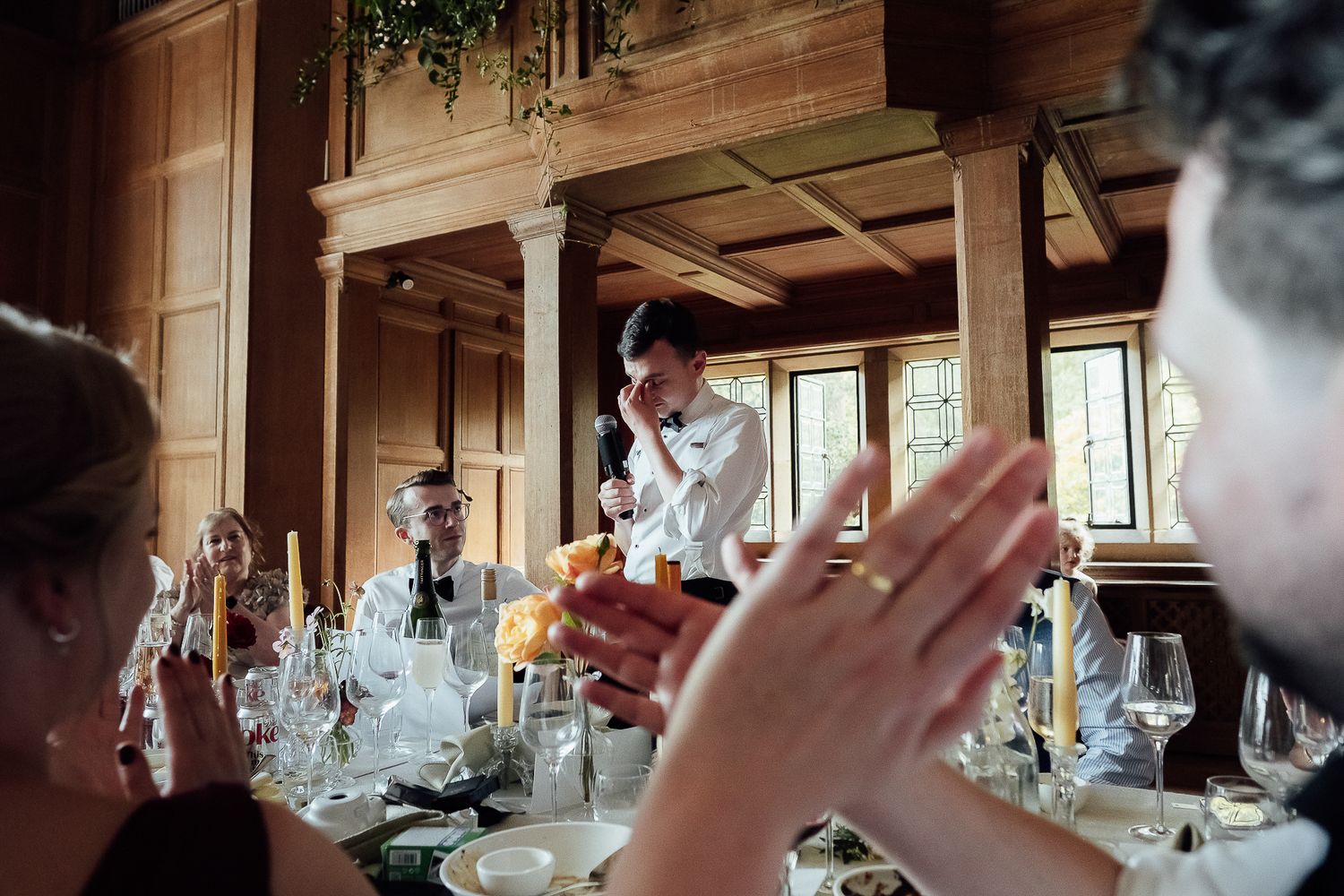 Guests applaud a speaker at an elegant formal dinner in a wood-paneled dining room with candlelit tables.