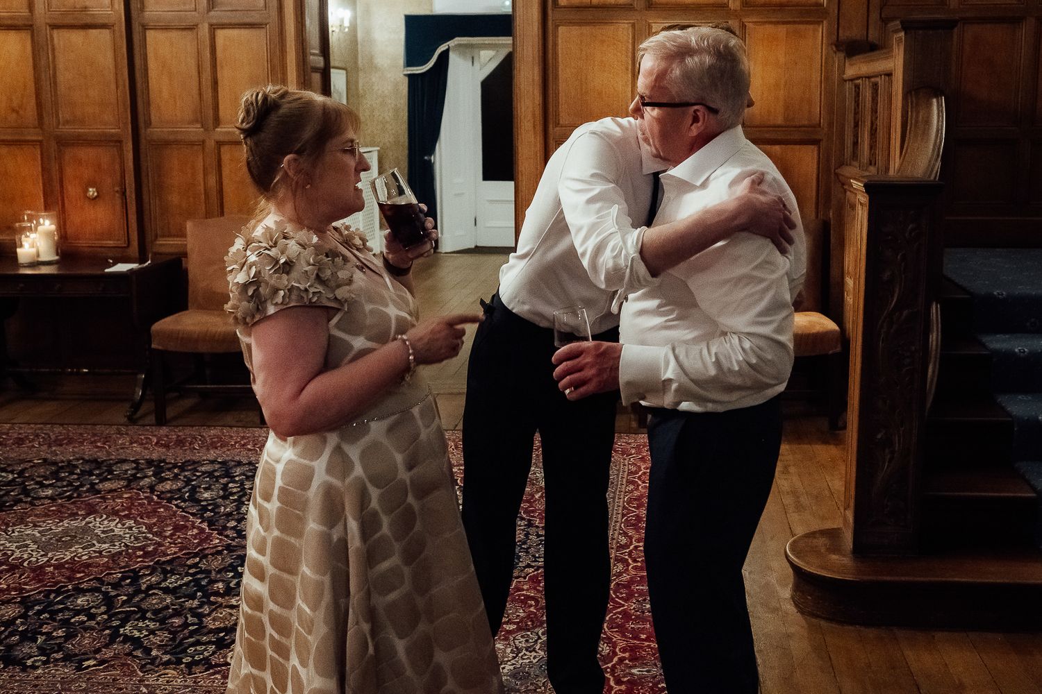Two people share a warm hug at an elegant wood-paneled reception hall while a woman in a floral dress looks on.