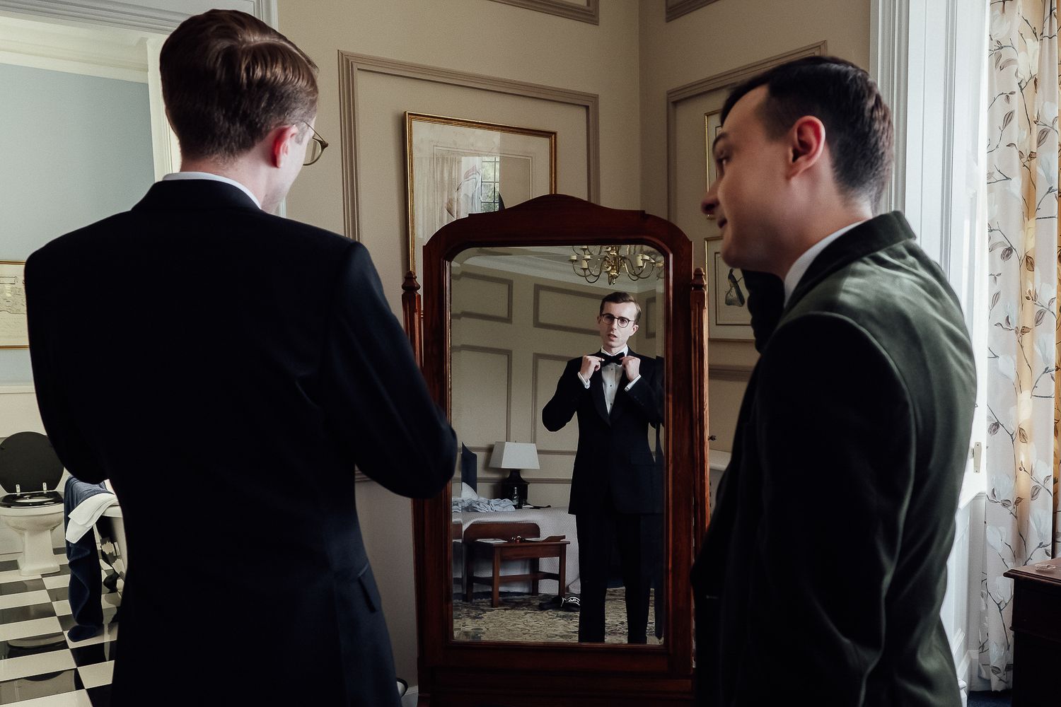 Two men in formal attire, one adjusting bow tie in ornate mirror, elegant room setting.