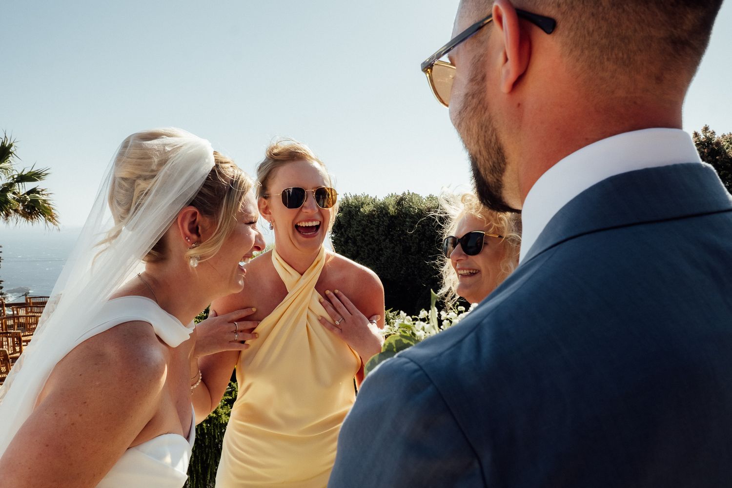 Bride and bridesmaids laughing joyfully with groom at outdoor wedding ceremony on sunny day.