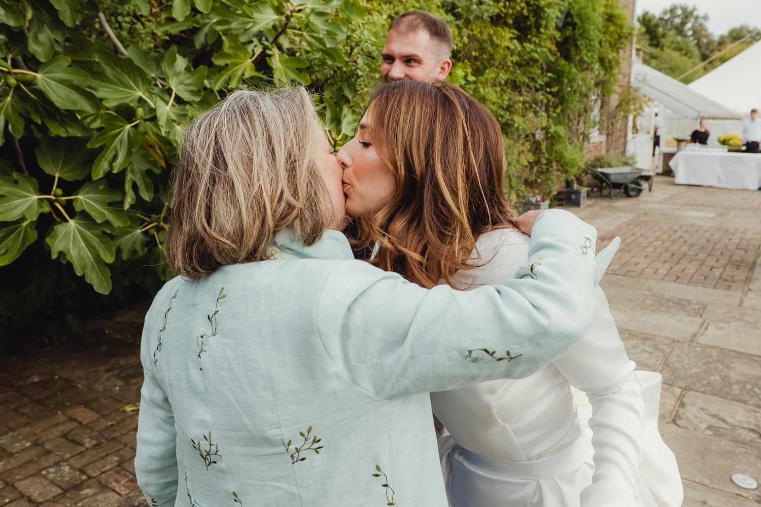 Two people in light colored clothing embrace while sitting outdoors on a stone path near green foliage.