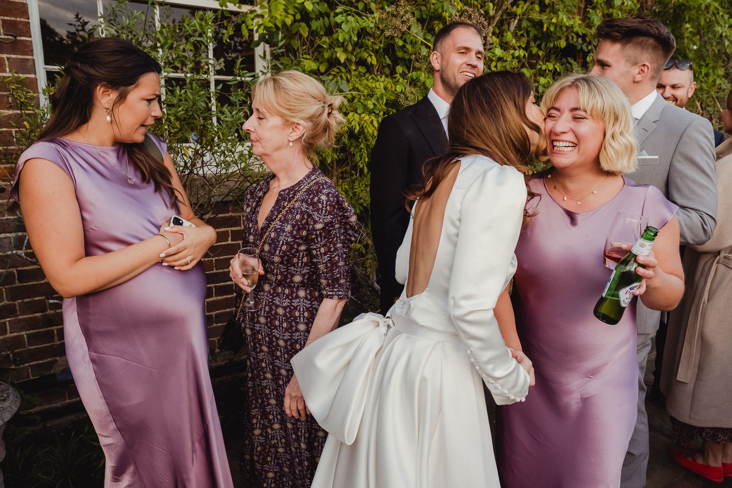 Wedding party members share joyful moments together in a garden setting wearing purple and white dresses.