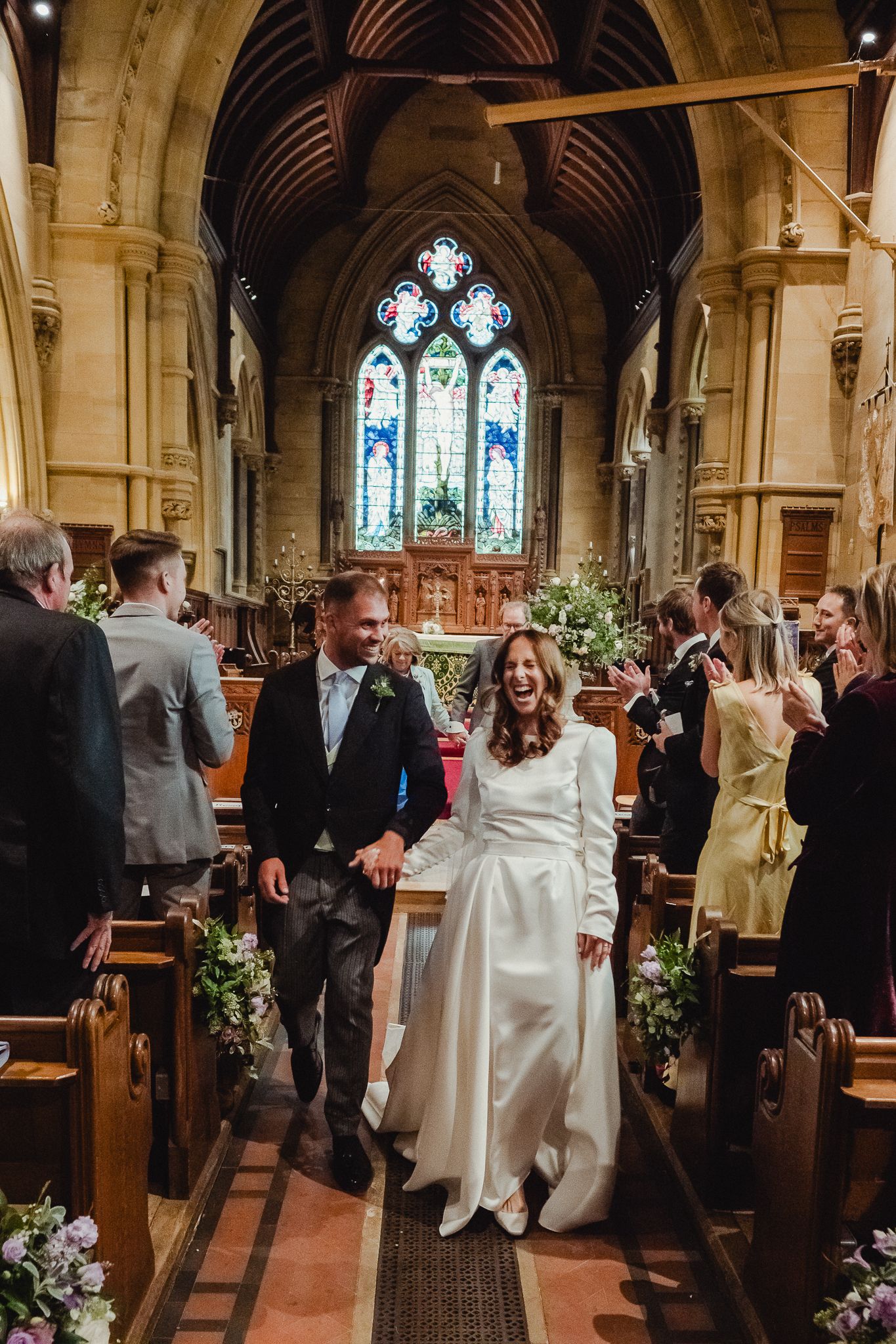 A wedding ceremony takes place inside a beautiful stone church with stained glass windows and floral decorations.