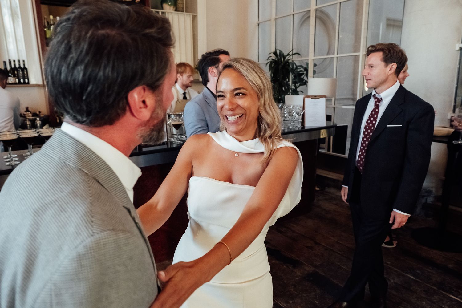 Smiling woman in a white dress embraces a man in a gray blazer at an elegant indoor reception, with guests and a man in a suit behind them.