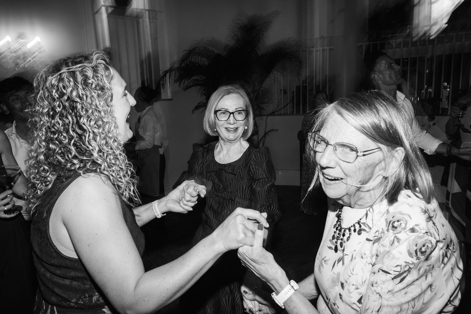 Three women in a black and white photo dance and hold hands at a lively event, smiling with joy as they celebrate together.