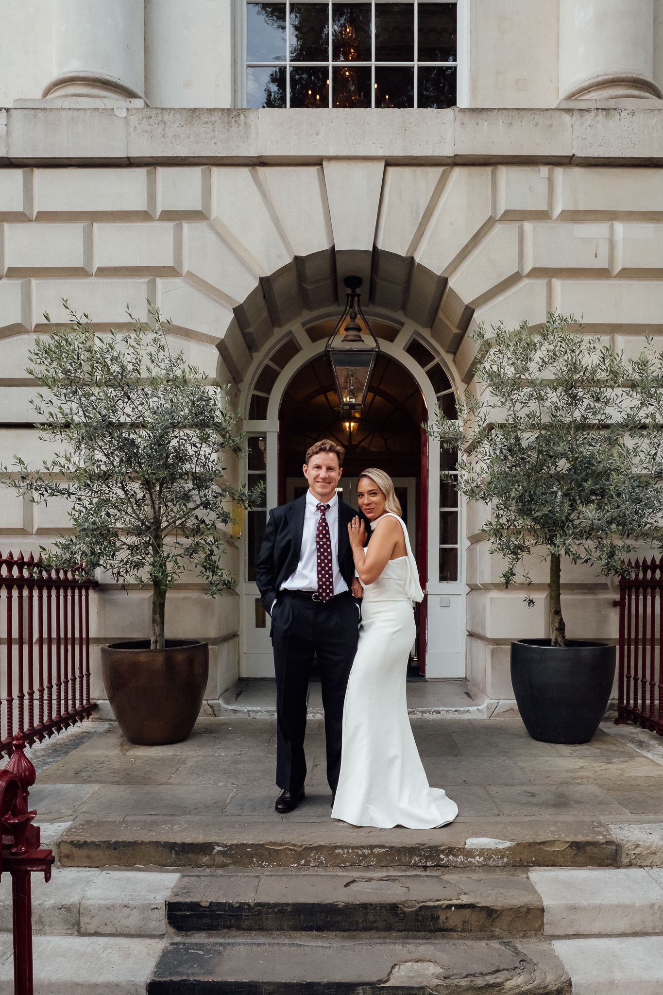 Bride in a white gown and groom in a dark suit embrace on stone steps beneath an arched doorway flanked by potted olive trees.